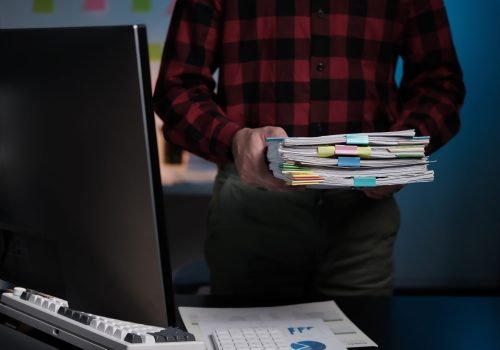 A man is holding a stack of papers in front of a computer monitor. The papers are organized and labeled with different colors
